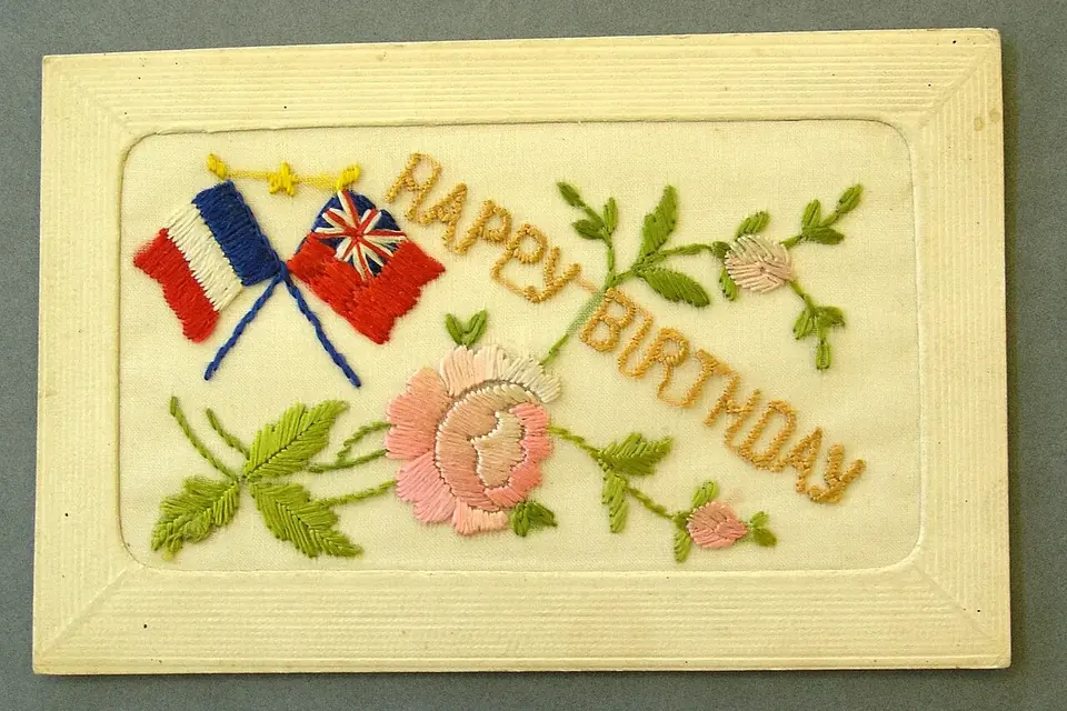 Carte postale ancienne brodée sur soie d'une rose, de feuillages et de deux drapeaux croisés, dans un encadrement de carton gaufré