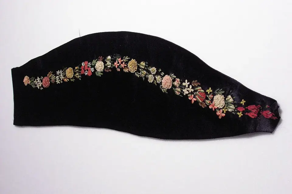 Pièce de tissu noir en forme de revers, brodée d'une guirlande de petites fleurs roses, jaunes et blanches au ruban